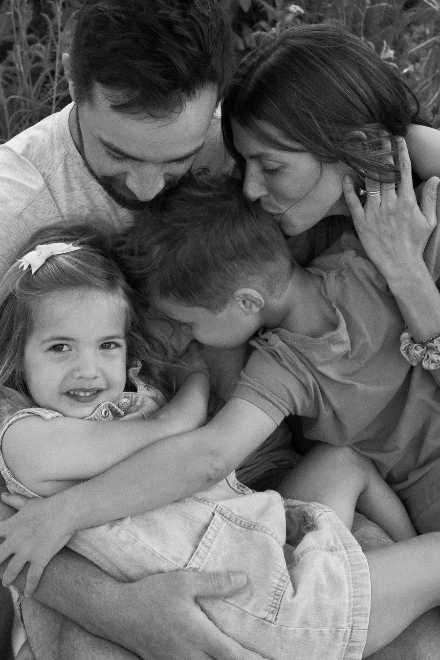 A family of four cuddled together in a black and white photo, mom kissing her son's head while her daughter grins at the camera