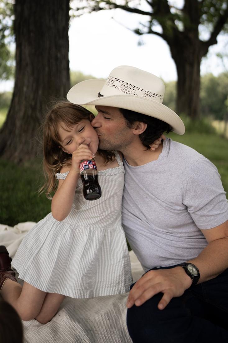 Dad in a cowboy hat kissing his daughter's cheek while she sips a soda