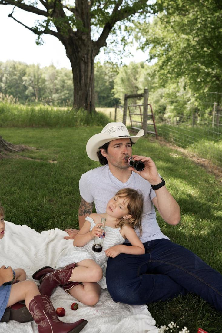 Dad in a cowboy hat drinking a soda on a picnic blanket while his daughter leans back against him