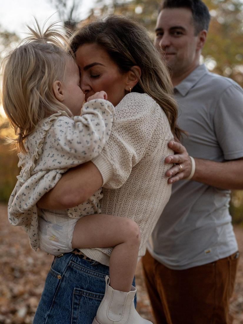 A mom pressing her forehead to her toddler’s while dad looks on, in the fall leaves