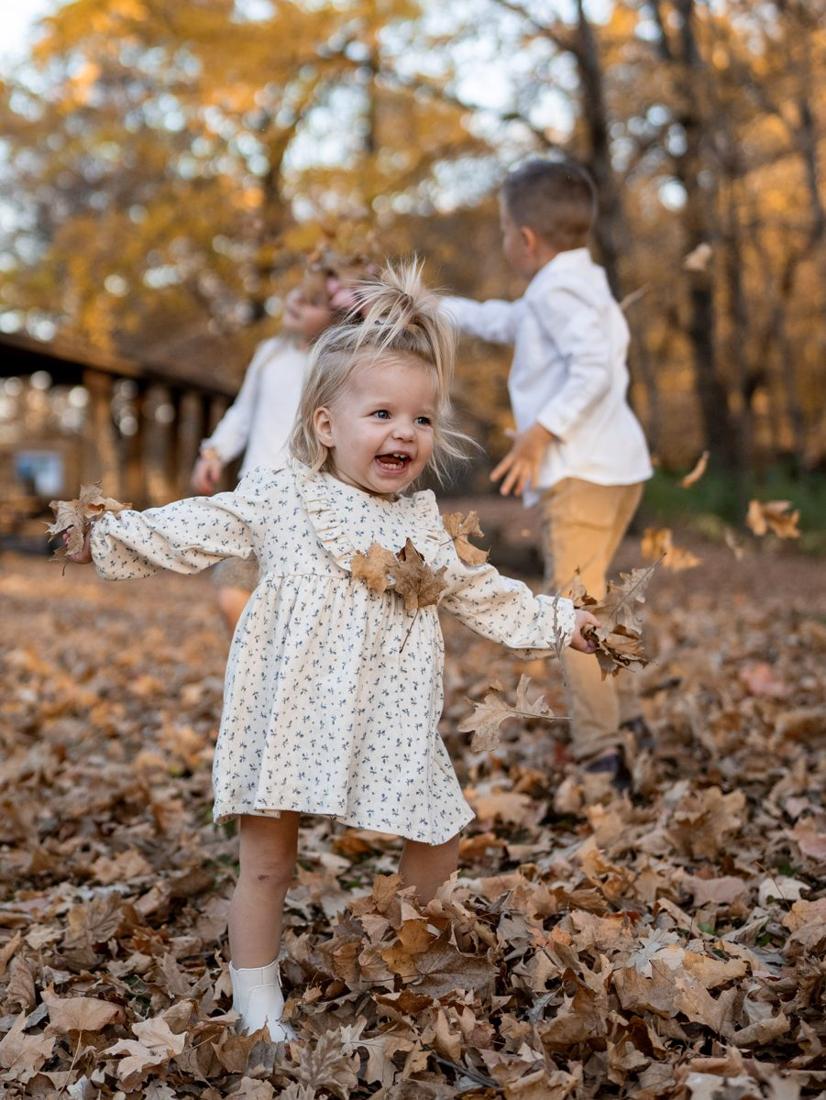A toddler giggling and throwing fall leaves while her brother and sister play behind her